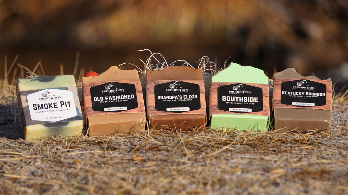 Five packaged artisanal tobacco products arranged in a row outdoors on dry grass, with labels reading Smoke Pit, Old Fashioned, Grandpa's Elixir, Southside, and Kentucky Bourbon. The packages are in different colors including green, tan, brown, and light green, set against a blurred natural background.