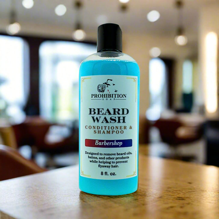 Prohibition Soap Barbershop Beard Wash conditioner and shampoo in clear 8 fl oz bottle with blue liquid on wooden table in authentic barbershop setting with warm lighting and blurred barber chairs in background