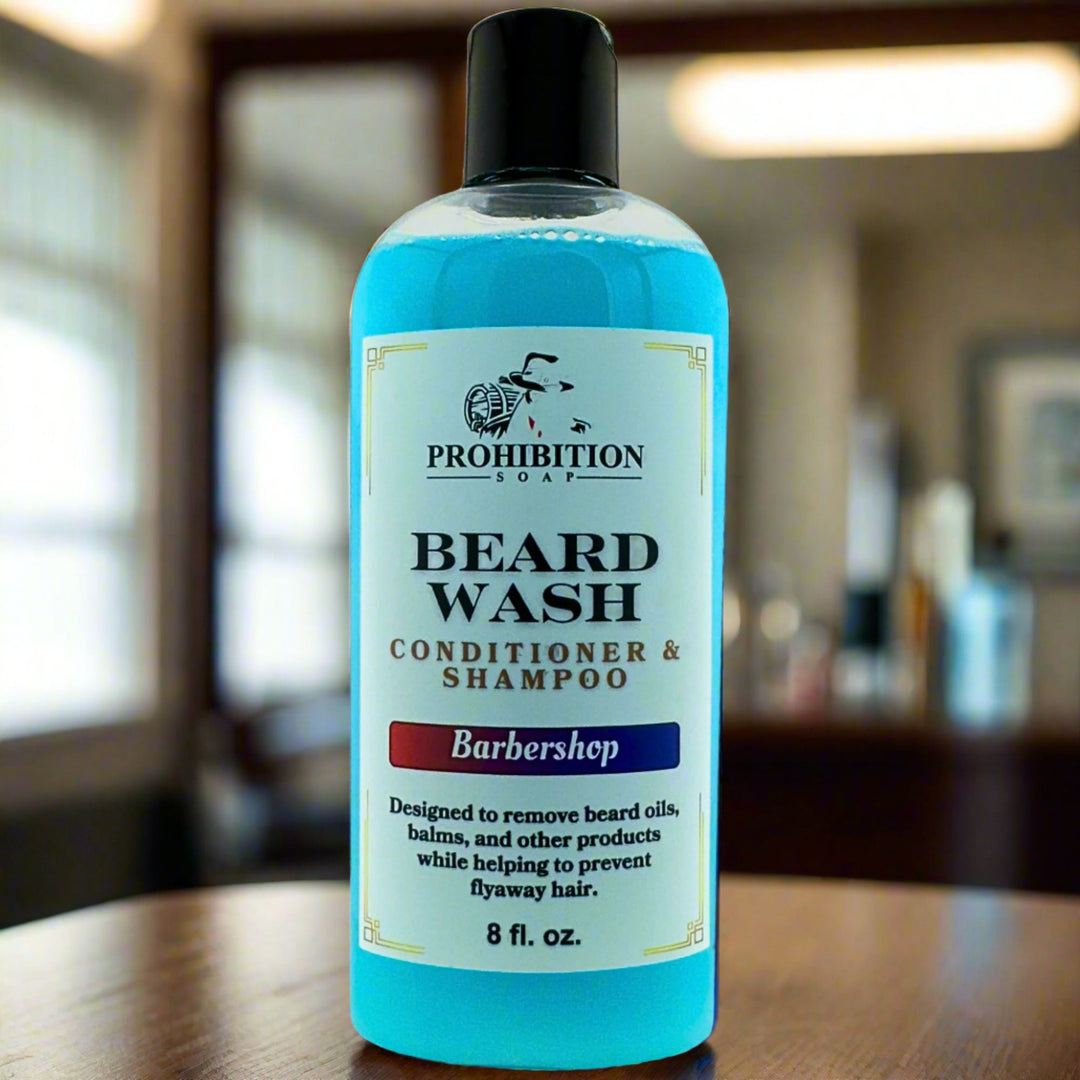 Prohibition Soap Barbershop Beard Wash conditioner and shampoo - clear 8 fl oz bottle containing blue wash liquid on wooden surface with blurred barbershop background, designed to remove beard oils and balms while preventing flyaway hair