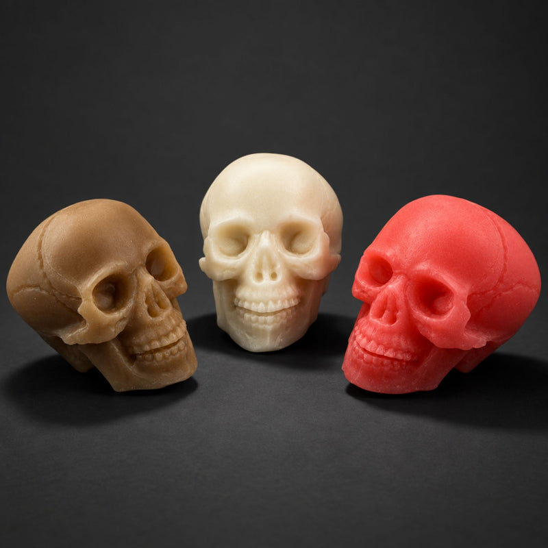 Three skull-shaped soaps in tan, ivory, and coral red, arranged on a dark gray background.