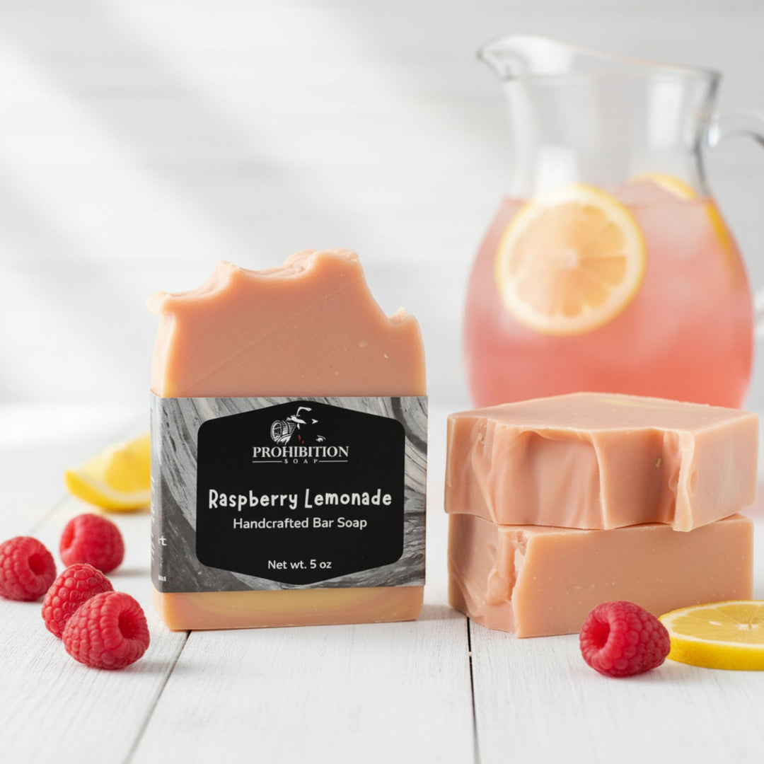 A bar of Prohibition Raspberry Lemonade handcrafted soap stands in front of two more bars, styled with fresh raspberries, lemon slices, and a pitcher of pink lemonade on a white wooden surface.