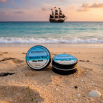 Two containers of Prohibition Mustache Wax on a beach with a pirate ship in the background.