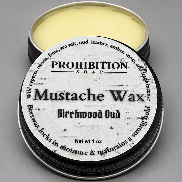 An open round metal tin of Prohibition Soap Mustache Wax in Birchwood Oud scent, showing the pale yellow wax inside, with the lid placed in front displaying the product label.