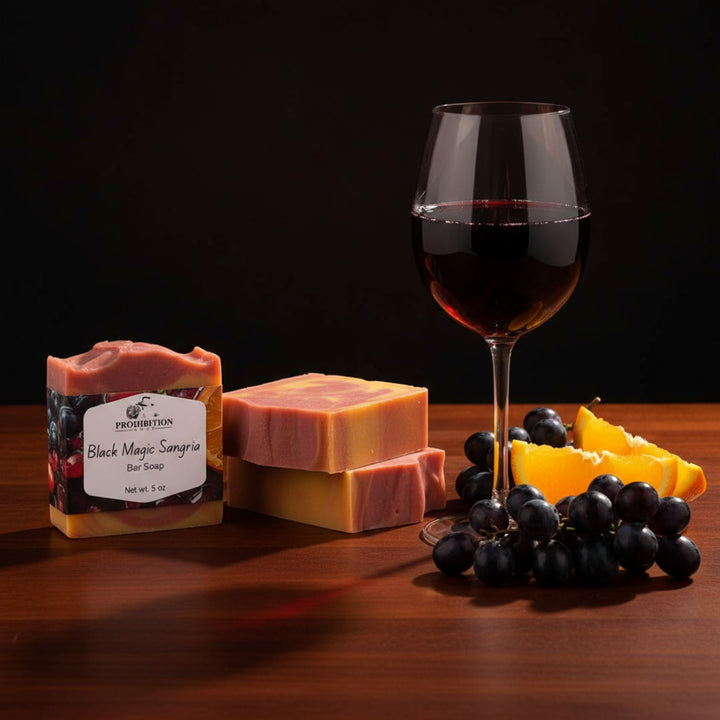 Three bars of soap, a glass of red wine, and grapes on a wooden surface with a dark background.