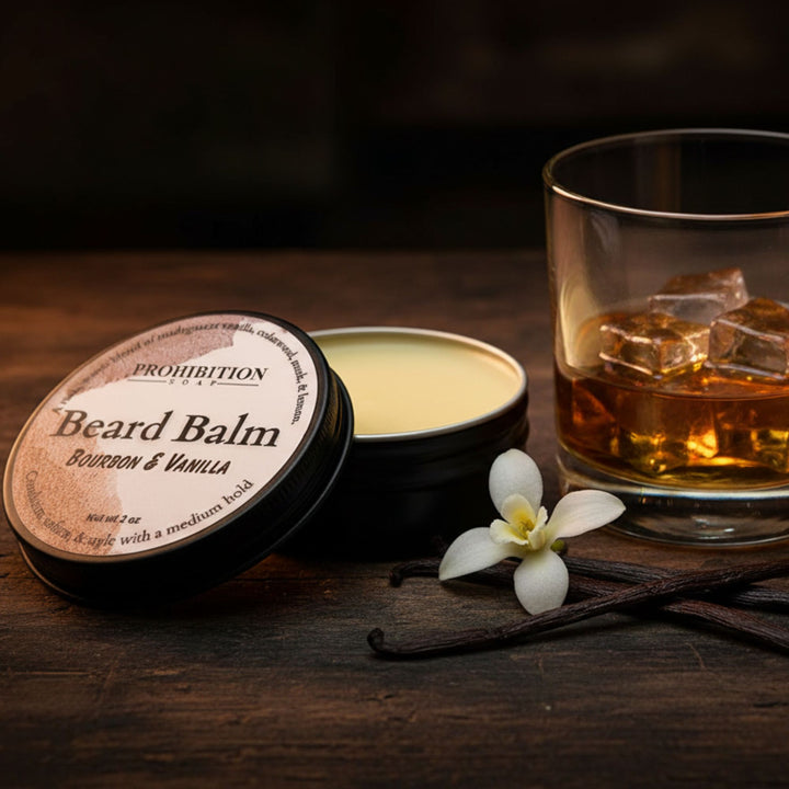 An open round tin of Prohibition Beard Balm in Bourbon &amp; Vanilla scent sits on a dark wooden table next to a glass of amber liquor with ice cubes, a vanilla bean, and a white flower. The lid is placed beside the open tin, showing the cream-colored balm inside.