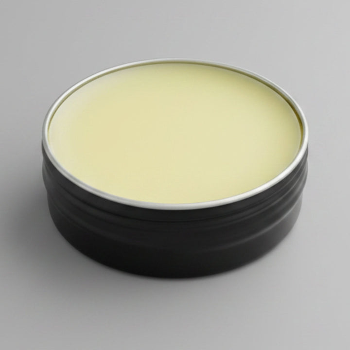 A round black metal tin with a pale yellow balm visible inside, photographed on a smooth light gray background.