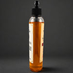 A clear plastic bottle filled with amber-colored soap, featuring a black dropper-style cap and cream-colored labels, set against a dark gray background.