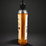A clear plastic pump bottle filled with amber-colored soap, standing upright against a dark gray background. The bottle has a black ribbed pump cap and a cream-colored label with black text.