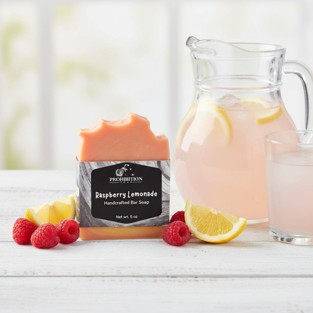 A bar of Prohibition Soap Raspberry Lemonade handcrafted bar soap stands upright on a white wooden surface, styled with fresh raspberries, lemon slices, a glass of pink lemonade, and a pitcher of pink lemonade in the background.