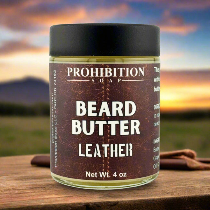 Prohibition Soap Beard Butter in Leather scent - 4 oz glass jar on rustic wooden surface with leather accessories and dramatic sunset sky background, showcasing rich brown leather-textured label design
