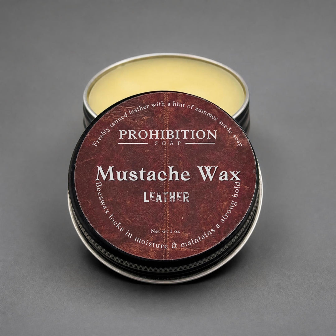 A round metal tin of Prohibition Soap Mustache Wax for leather, with the lid placed on top of the open tin showing the pale yellow wax inside, set against a dark gray background.
