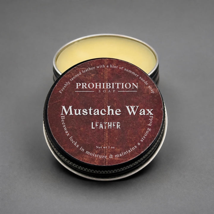 A round metal tin of Prohibition Soap Mustache Wax for leather, with the lid placed on top of the open tin showing the pale yellow wax inside, set against a dark gray background.