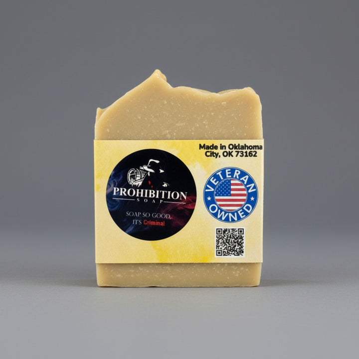 A single bar of handmade soap in a rustic, hand-shaped block, featuring a pale yellow-beige color. It has a yellow paper label with a black circular logo for &