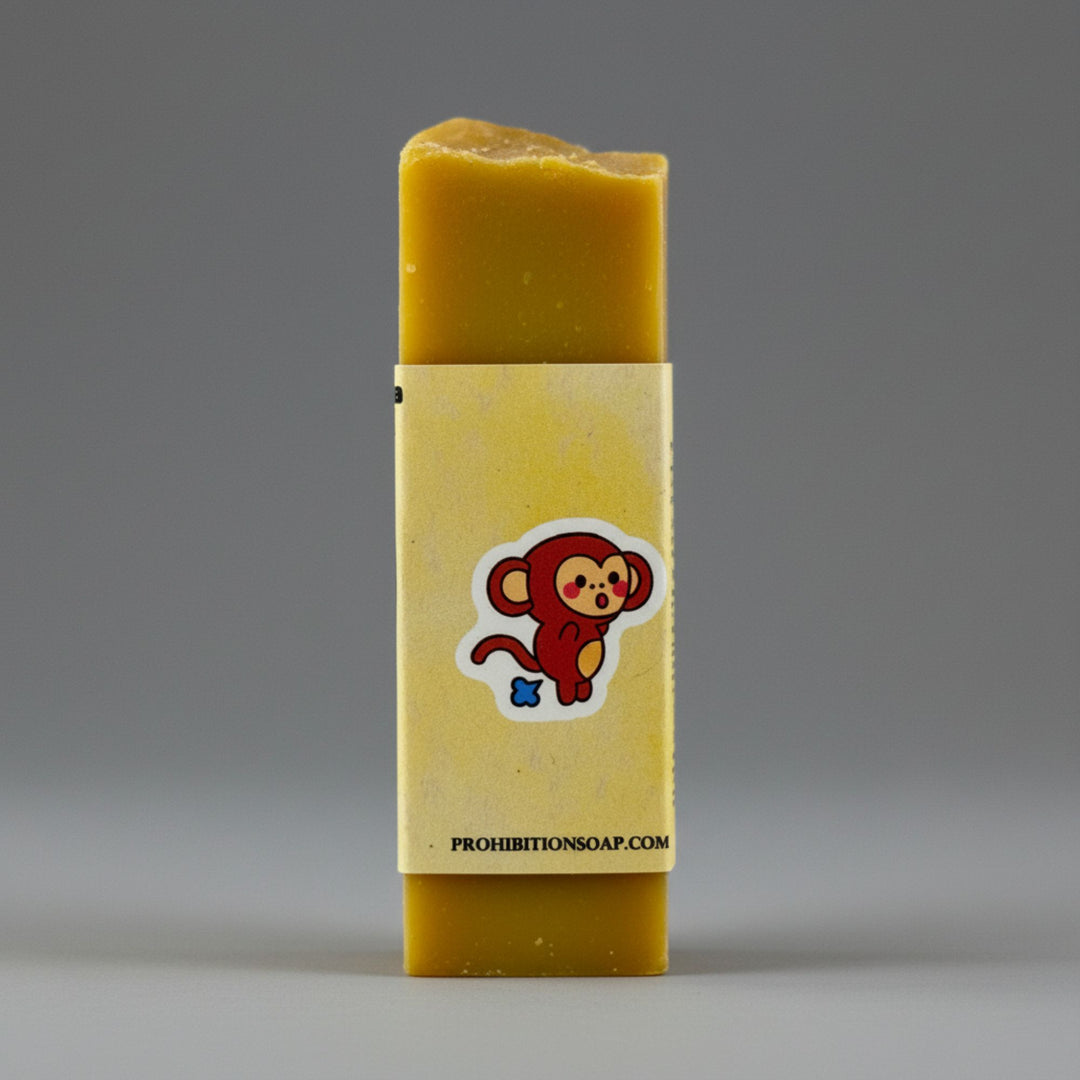 A rectangular bar of yellow soap standing upright, wrapped in a pale yellow paper sleeve with a cartoon monkey sticker and the text &