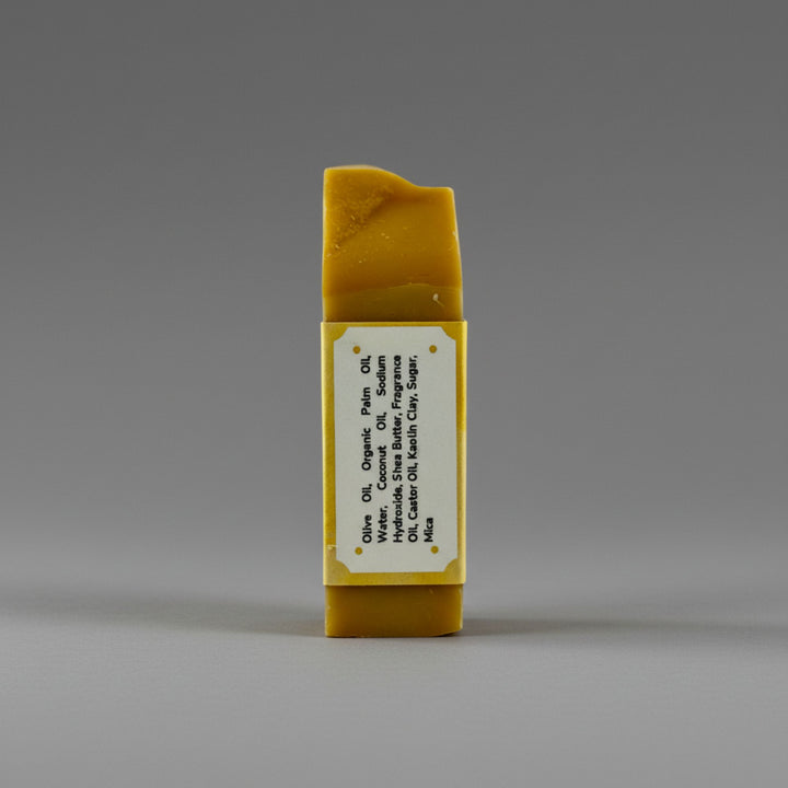 A rectangular bar of handmade soap in a warm banana yellow color, standing upright on a neutral gray background. It features a white paper label wrapped around the center with black text detailing its ingredients: olive oil, coconut oil, shea butter, and clay powder.