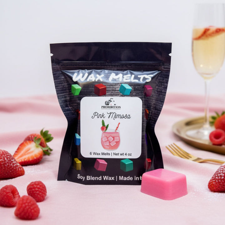 Packaged wax melts with a pink mimosa design on a pink surface with strawberries and a glass of champagne.