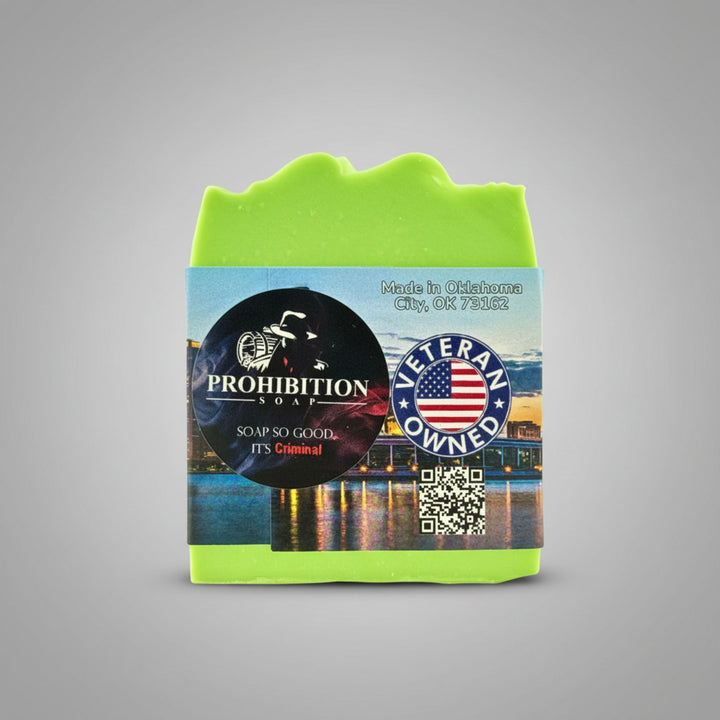 A single bar of bright lime green soap with a decorative wavy top edge, wrapped in a colorful paper label. The label features a black circular logo for &