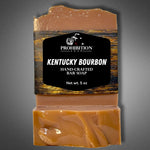 Kentucky Bourbon bar soap with Prohibition Soap label standing beside two unwrapped bars stacked to show the swirled amber interior, on a gray background