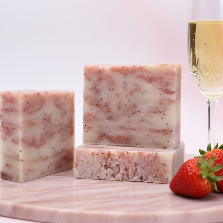 Three bars of strawberry-flavored soap on a marble surface with strawberries and a glass of champagne in the background.