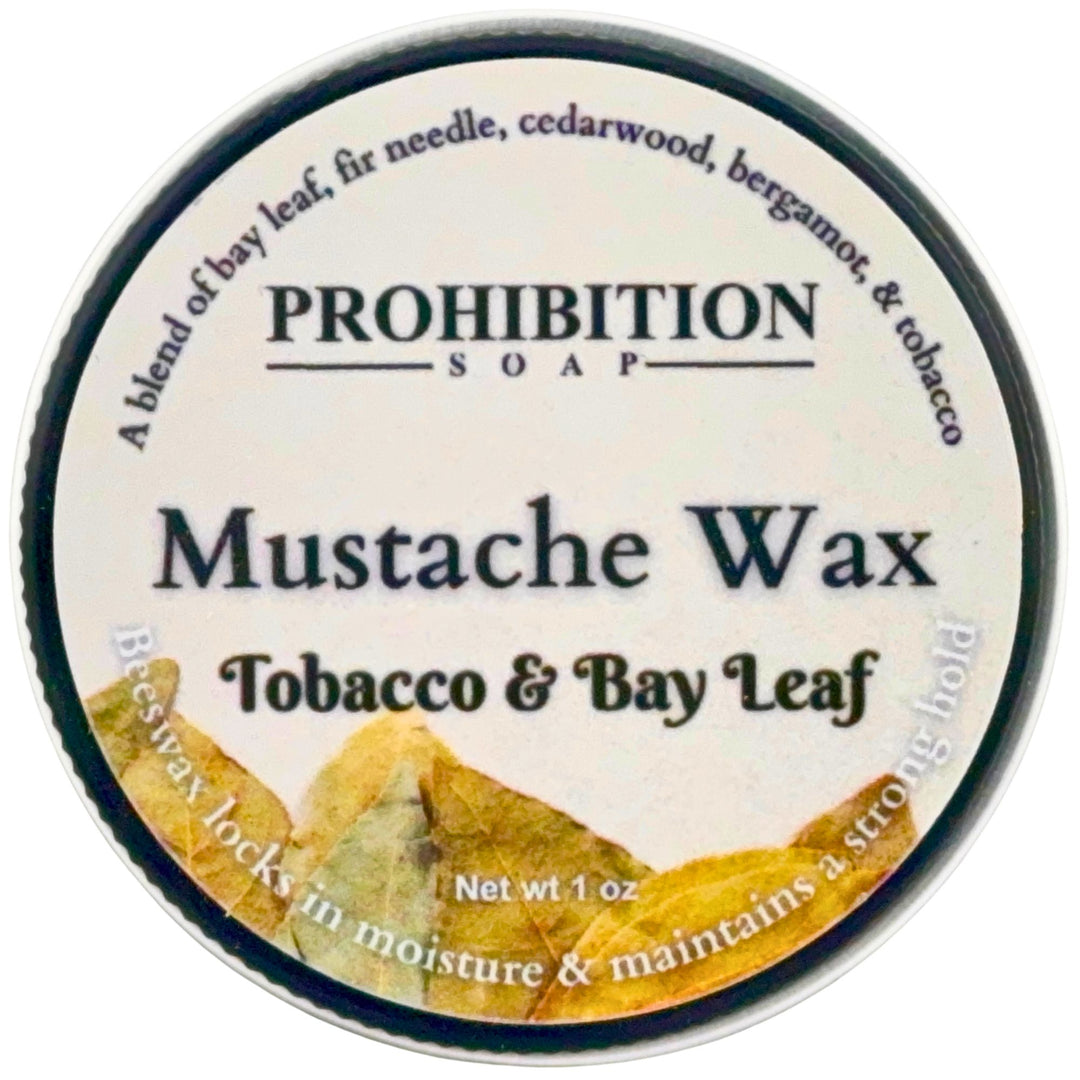 A round tin of Prohibition Soap Mustache Wax in Tobacco & Bay Leaf scent, featuring a white label with black text and a graphic of yellow-brown leaves at the bottom.