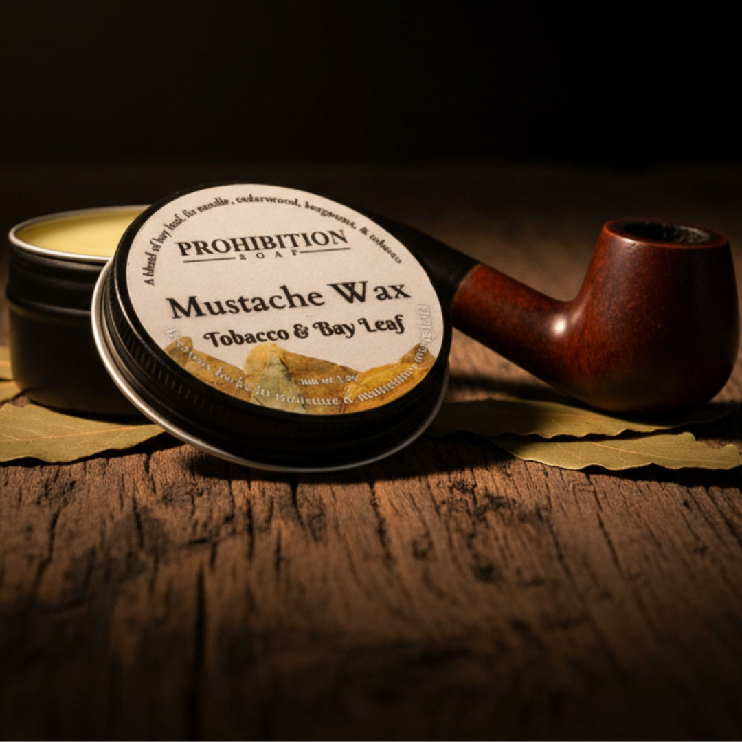 A tin of Prohibition Mustache Wax with its lid off, placed on a rustic wooden surface next to a dark brown smoking pipe. The wax itself is visible in the open tin, and the label reads &