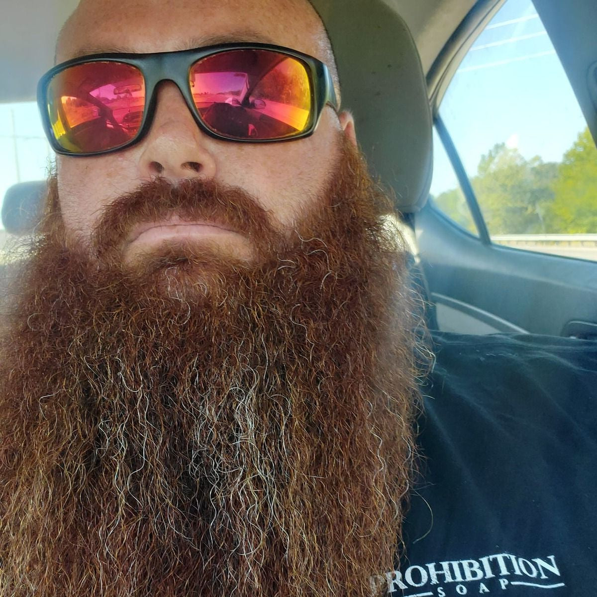 A close-up selfie of a man with a long brown beard wearing black sunglasses with red mirrored lenses and a black t-shirt inside a car.