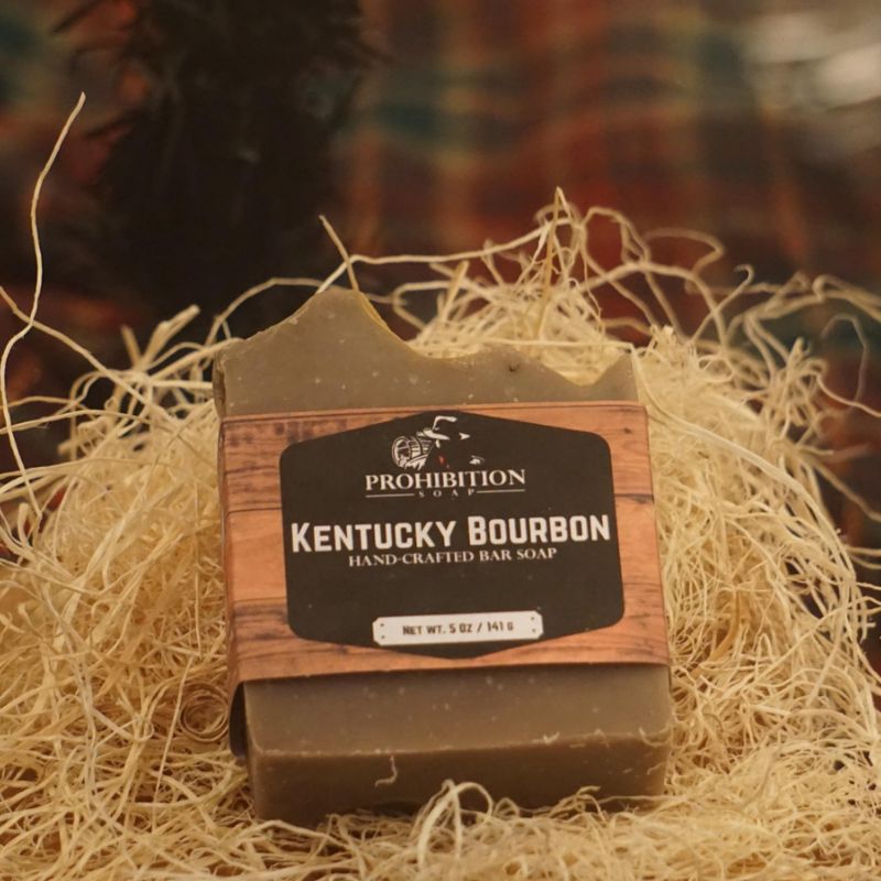Kentucky Bourbon bar soap nestled in a bed of wood wool shavings, with a warm plaid background suggesting a rustic, handcrafted feel