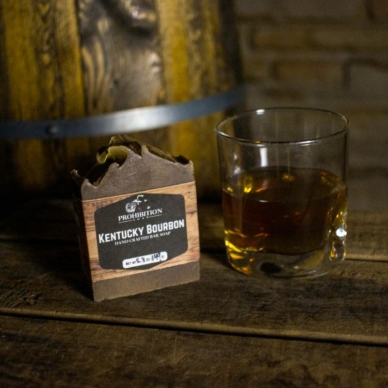 Kentucky Bourbon bar soap beside a rocks glass of bourbon, resting on weathered wood planks in front of an oak barrel